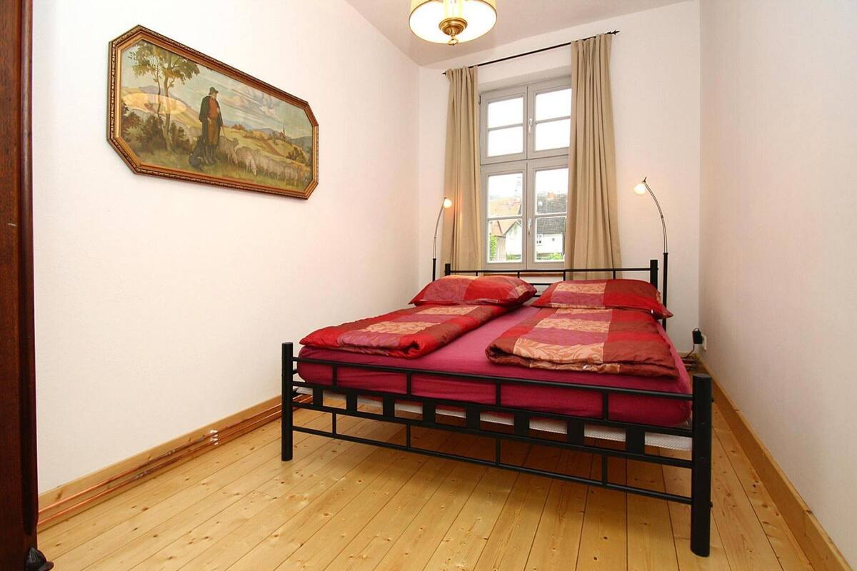 A bedroom with a wooden floor, a bed with red bedding, a window with curtains, and a painting on the wall.