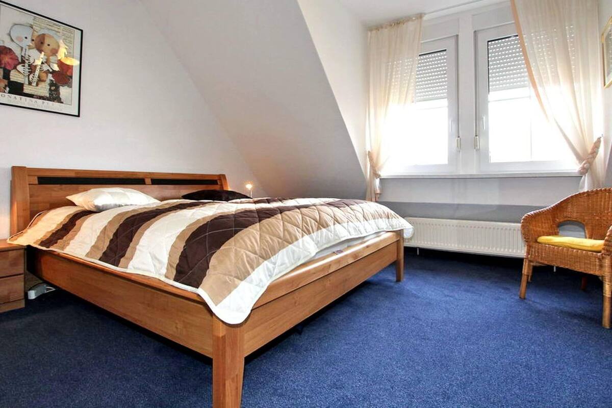 Une chambre à coucher avec un lit en bois, une moquette bleue, un fauteuil en osier et une fenêtre avec des rideaux.