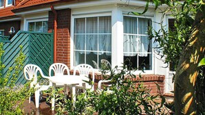 A two-story house with a red-tiled roof, white exterior walls, and a brick chimney. There is a patio with white plastic chairs and a table. - Reihenhaus, Dornumersiel (Dornum)