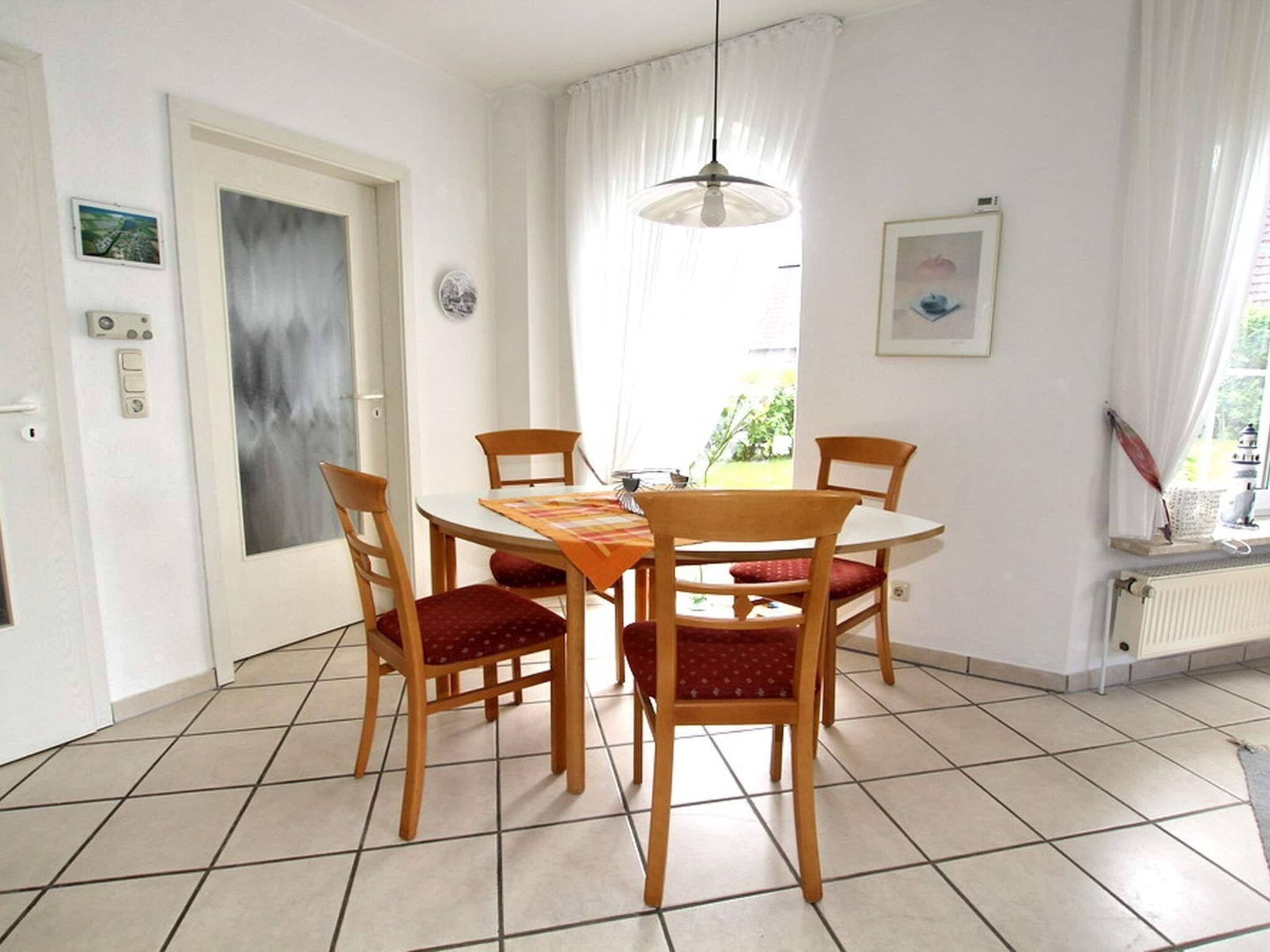 A dining area with a round table, four wooden chairs, a patterned tablecloth, a pendant light, and a window with curtains.