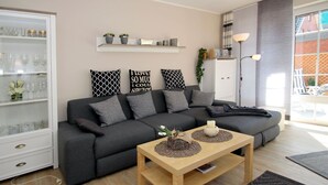 A modern living room with a grey sectional sofa, a wooden coffee table, and a glass cabinet with decorative items. - Apartment in a Quiet Location in Dornumersiel (Dornum)