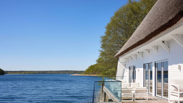 A wooden deck with a glass railing overlooking a calm body of water, a white building with large windows, and a thatched roof. - Semi-detached Houses at the Drewitzer See, Drewitz (Nossentiner Huette)