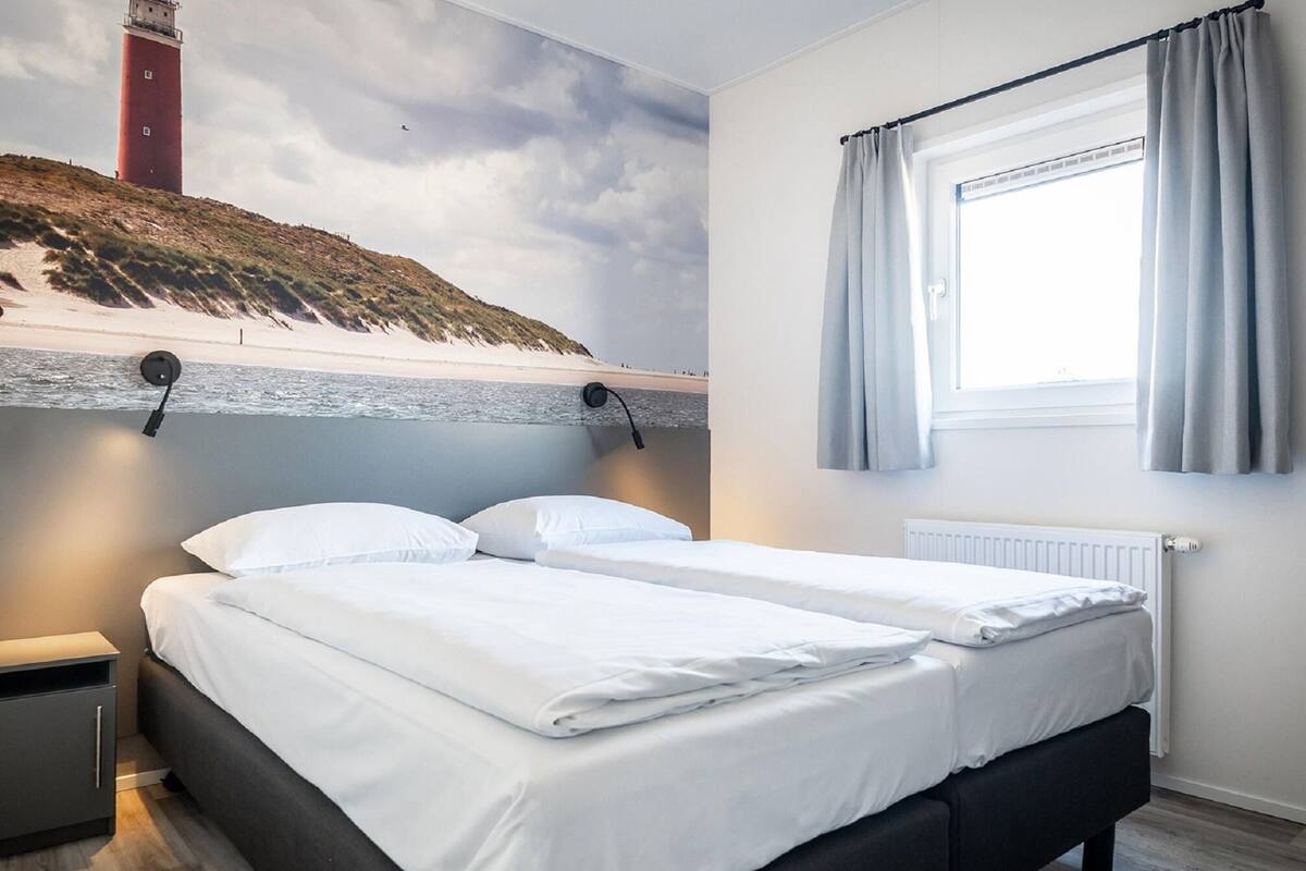 A bedroom with two beds, a nightstand, a window with curtains, and a mural of a lighthouse on the wall.