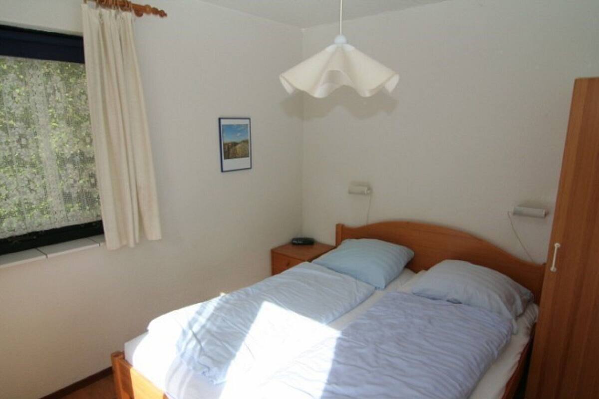 A bedroom with a wooden bed, white curtains, a small bedside table, a nightstand, a wall-mounted light, and a window with a view of trees.