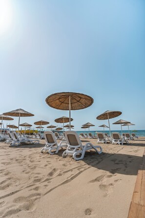 Une plage bordée de rangées de chaises longues blanches et d’ombrelles en chaume.