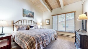 Un dormitorio con cama, dos mesitas de noche, cómoda, ventana con persianas, techo con vigas de madera y un cuadro en la pared.