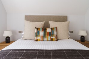 A neatly made bed with a striped white duvet, two beige pillows, and a colorful patchwork pillow. There are two bedside tables with lamps on either side of the bed.