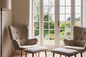 A room with two upholstered armchairs and a matching ottoman, a floor lamp, and a large window with a view of greenery.