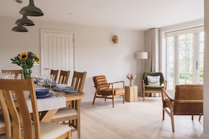 A dining area with a round table set for four, surrounded by wooden chairs. There is a side table with a lamp and a vase of flowers. The room has a large window with curtains, a door, and a wall-mounted light fixture.