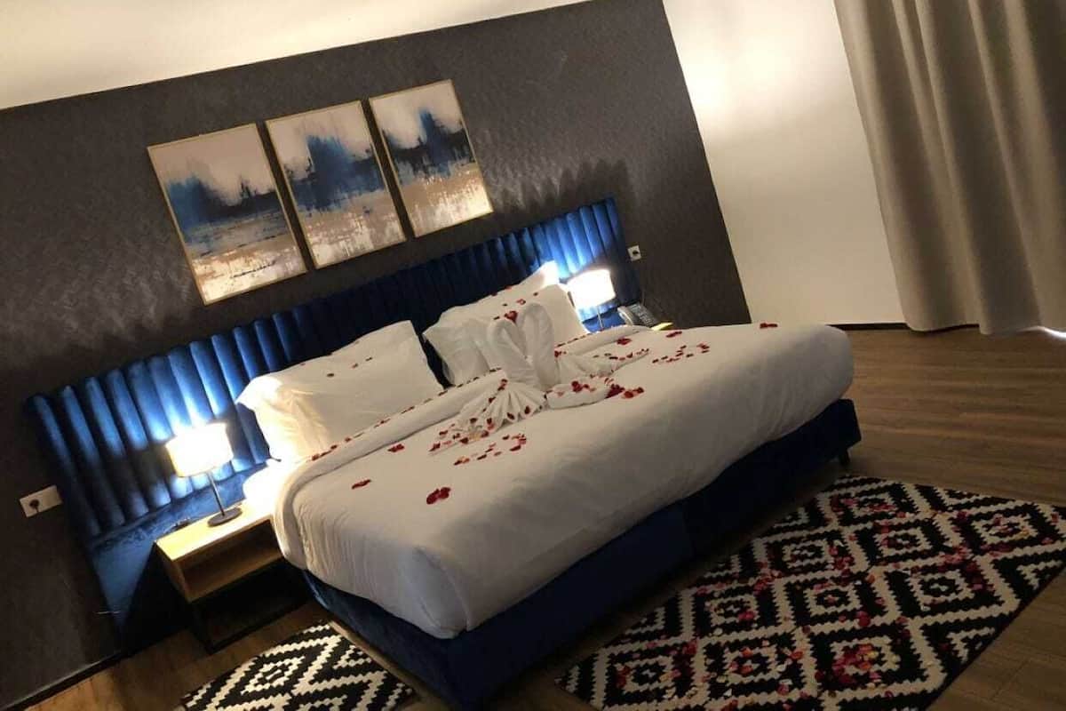A hotel room with a bed, a headboard, a nightstand, a lamp, a patterned rug, and artwork on the wall.