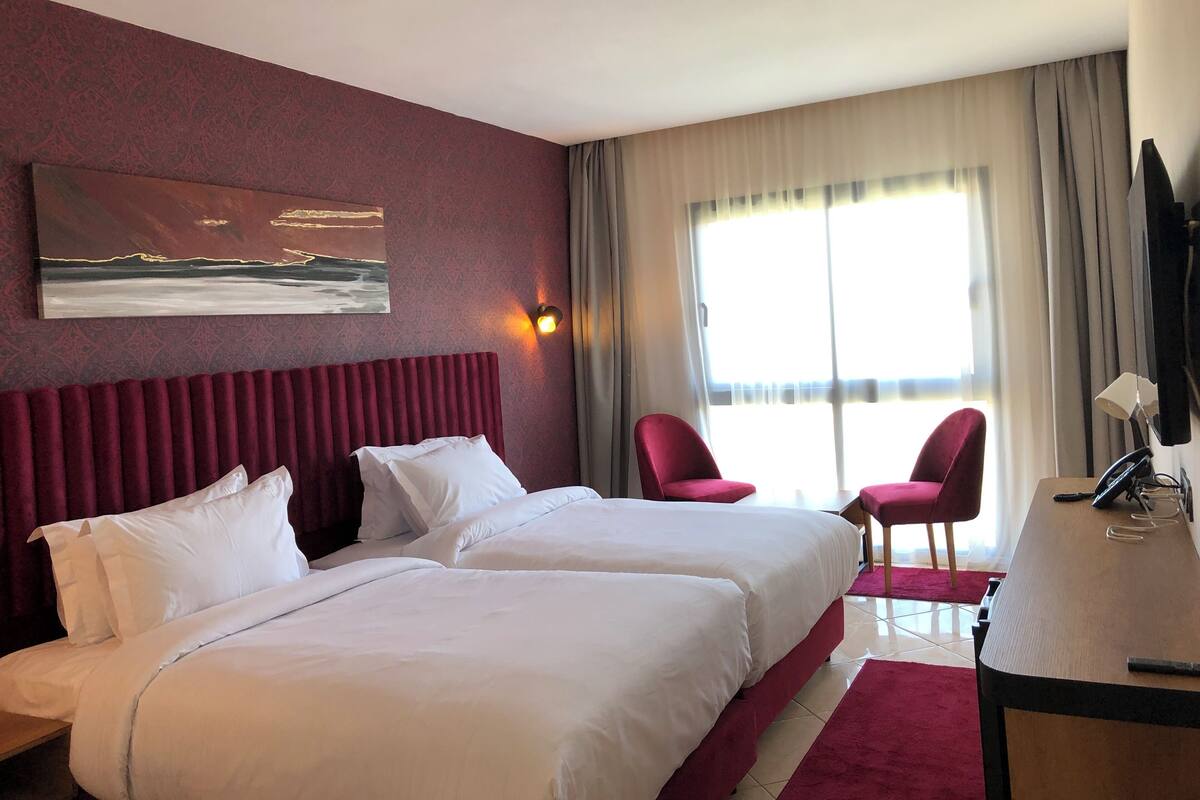 A hotel room with two beds, a painting, a window with curtains, and a desk with a lamp.