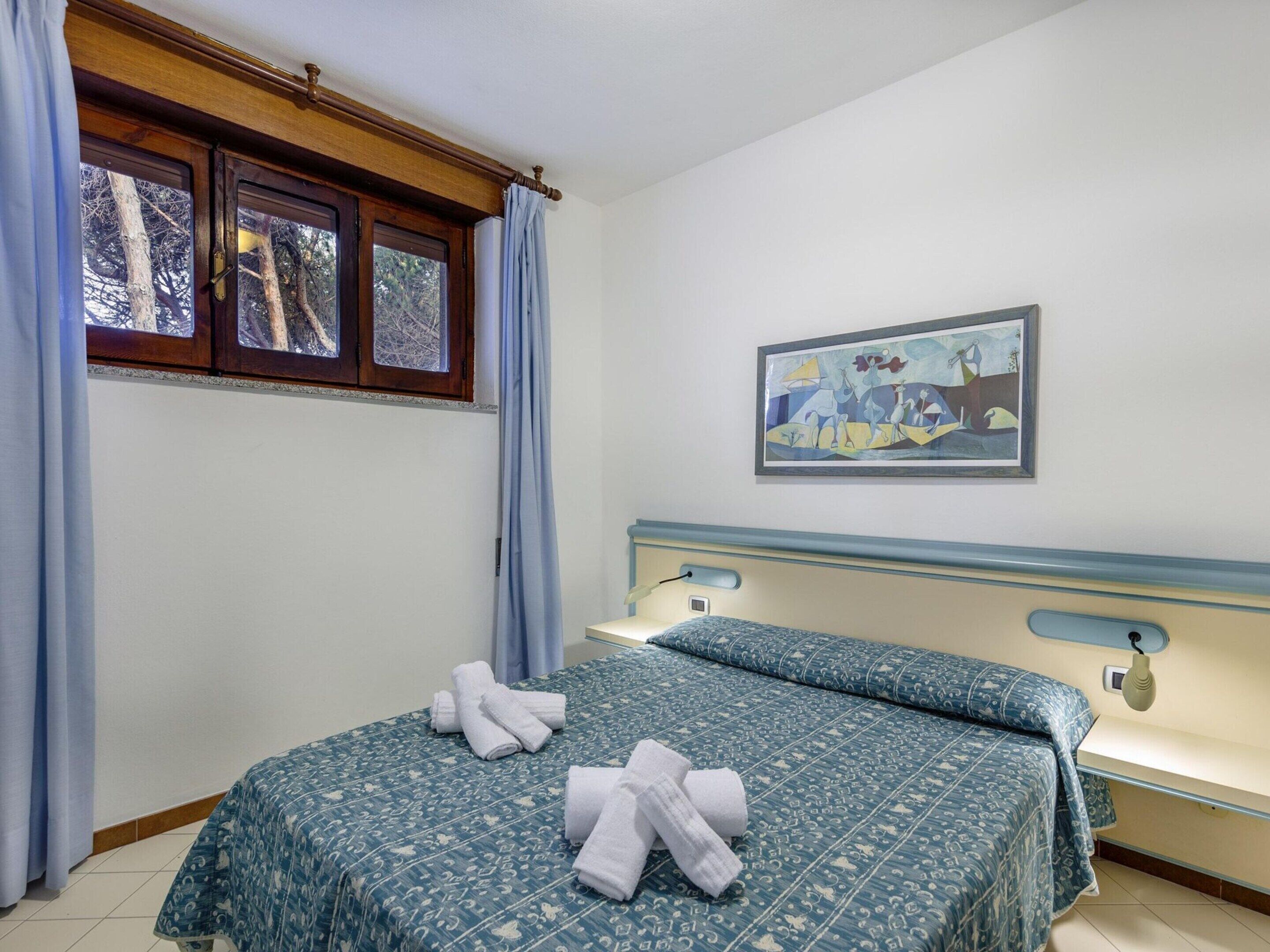 A hotel room with a bed, a window with curtains, a painting on the wall, and a bedside table with a lamp.