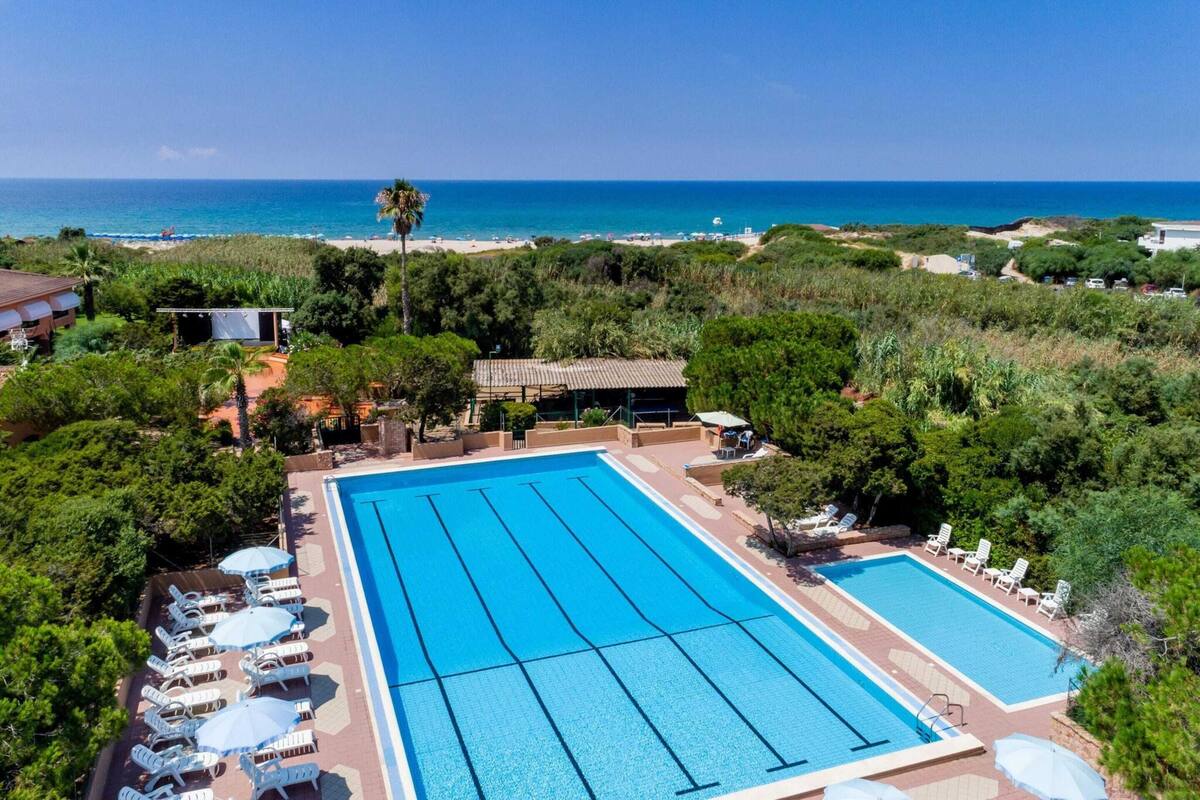 Uma grande piscina ao ar livre com várias pistas de natação, rodeada por espreguiçadeiras e guarda-sóis, com vista para uma praia e o oceano ao fundo.