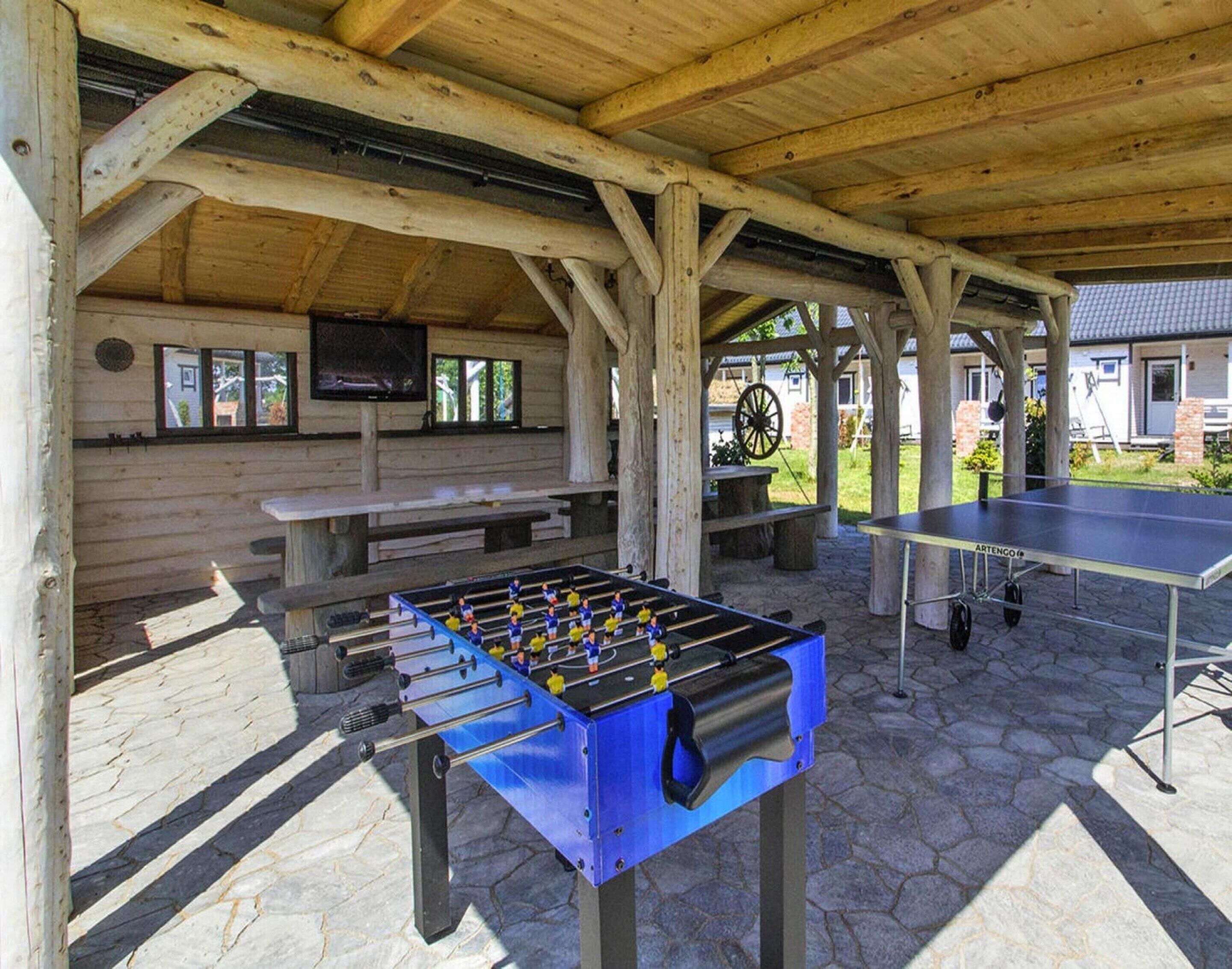 An outdoor recreational area with a foosball table, a ping pong table, and a covered seating area with a television.