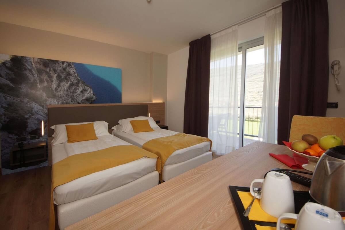 A hotel room with two beds, a wooden desk, a fruit bowl, and a painting on the wall.