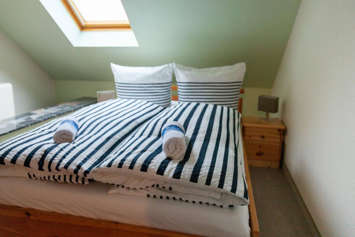 Een net opgemaakt bed met gestreept beddengoed en kussens, een houten nachtkastje met een lamp, en een dakraam erboven.