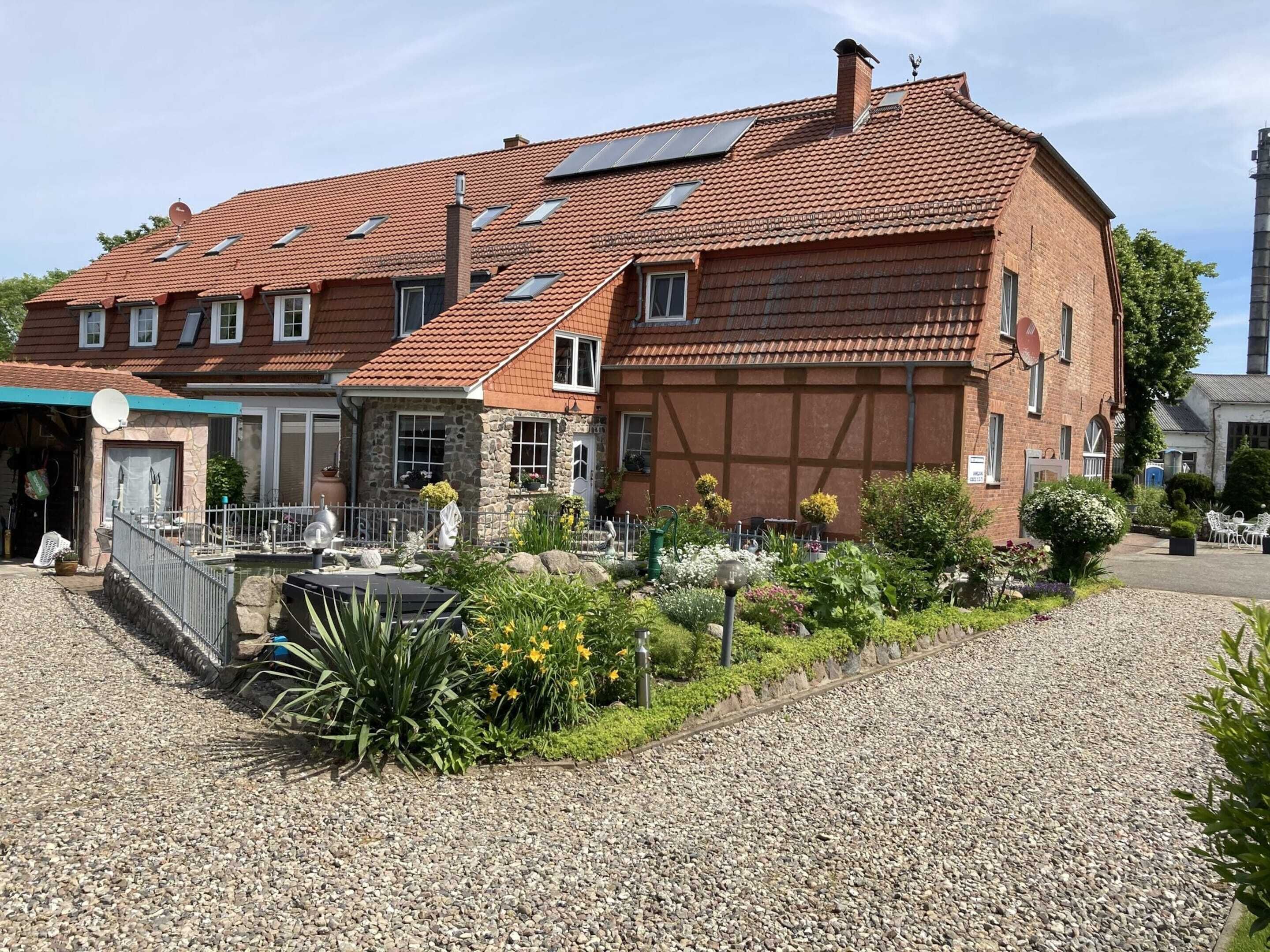 A large brick building with multiple windows, a gravel driveway, and a garden area with various plants and flowers.