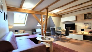 Un espace de vie moderne de style loft, doté d’un plafond haut, de poutres apparentes, d’un coin salon confortable avec un canapé et des fauteuils, d’une table à manger et d’un coin cuisine avec un téléviseur et des placards.