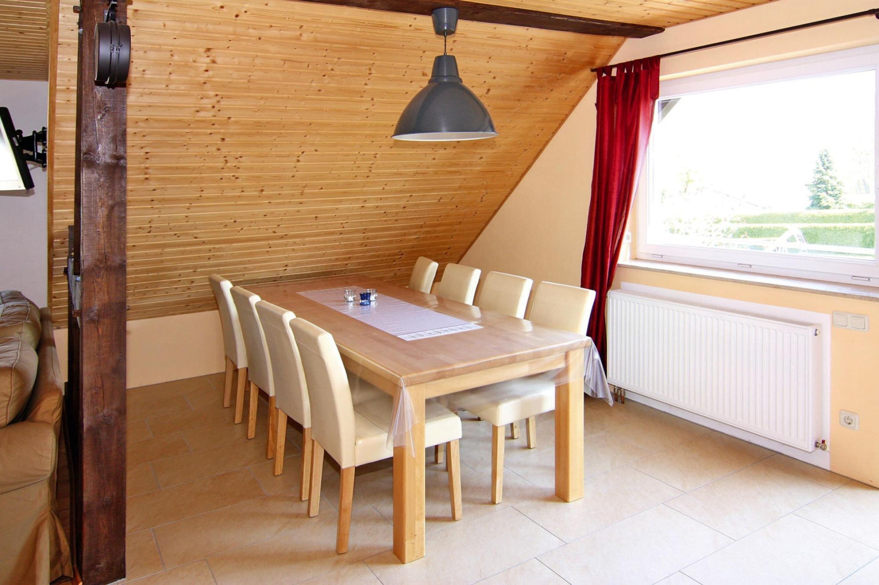 A dining area with a wooden table and chairs, a hanging light fixture, a window with curtains, and a radiator.