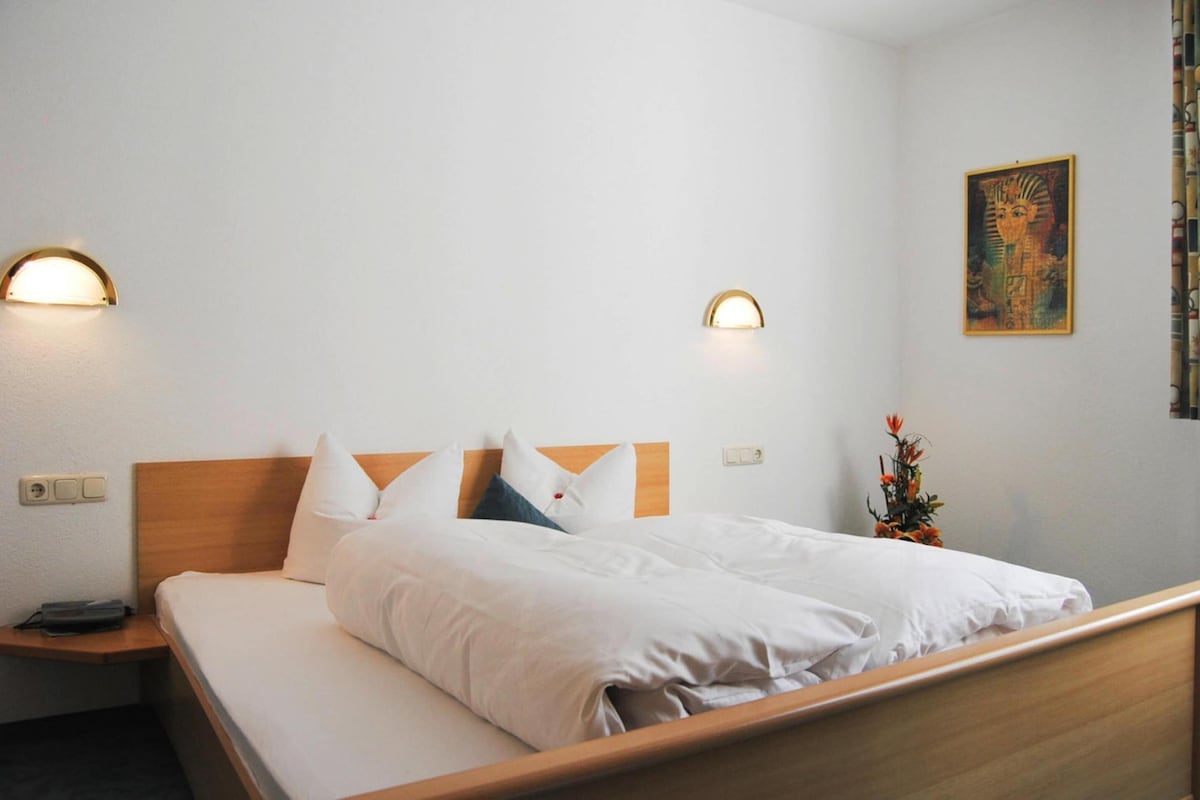 A hotel room with a wooden bed, white bedding, two wall-mounted lights, a nightstand, a power outlet, and a framed artwork on the wall.