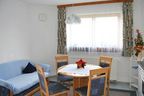 A room with a blue sofa, a wooden dining table with chairs, a window with curtains, a radiator, and a clock on the wall. - Apartments Home Val Sinestra, Kappl (Kappl)