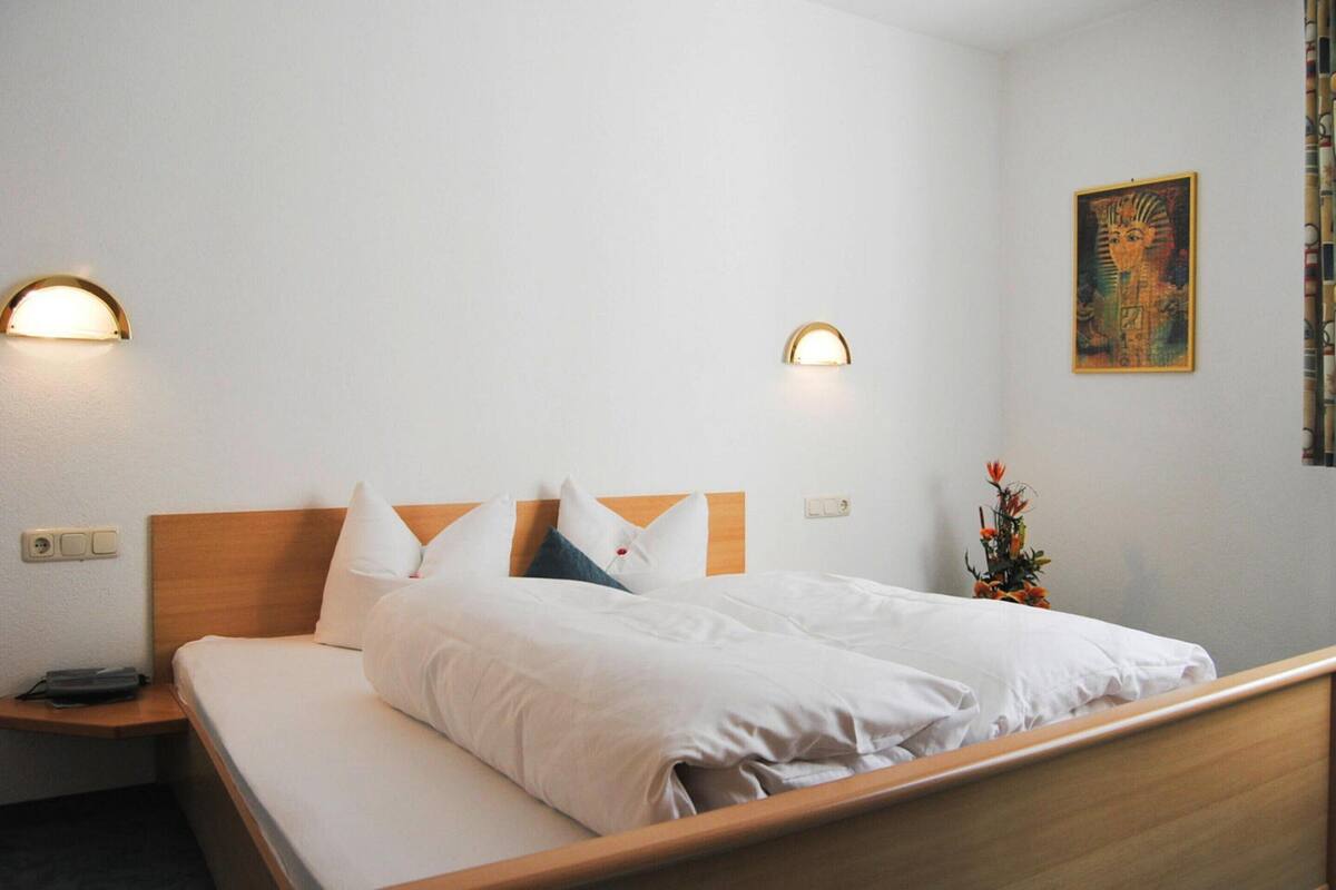 A neatly made bed with white linens and pillows, a wooden headboard, and two wall-mounted light fixtures.