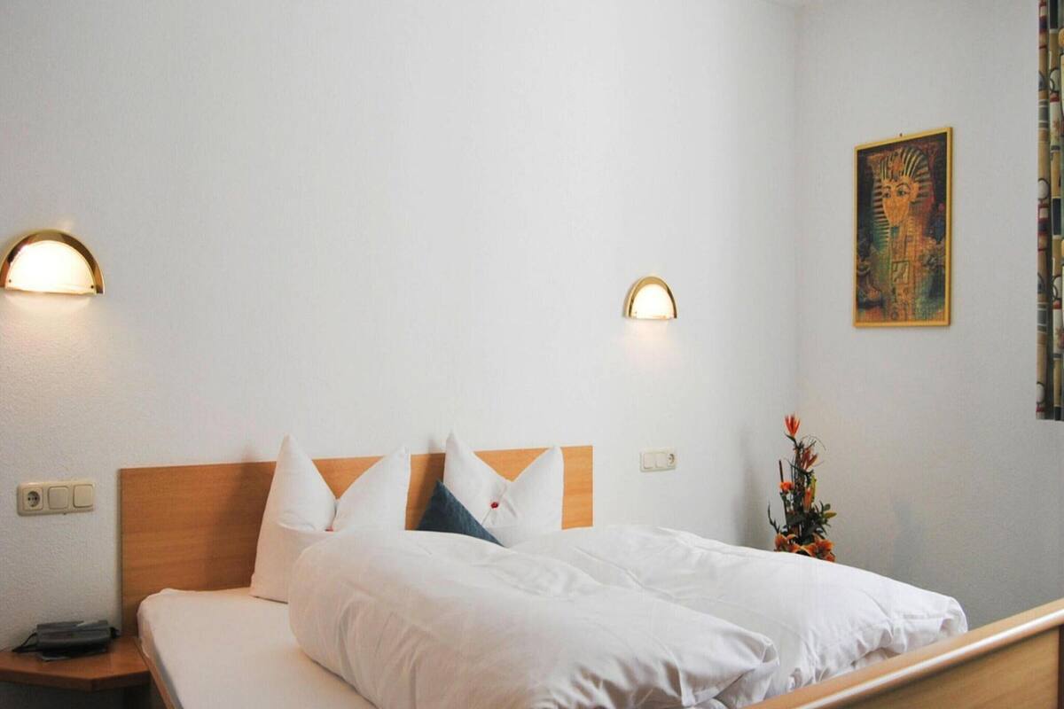 A neatly made bed with white linens and pillows, two bedside lamps, a wall-mounted light, and a painting of a lioness.
