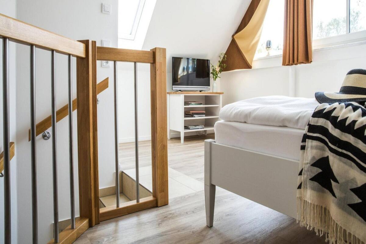 A modern bedroom with a sloped ceiling, a wooden staircase, a TV on a white stand, a bed with a patterned blanket, and a window with curtains.
