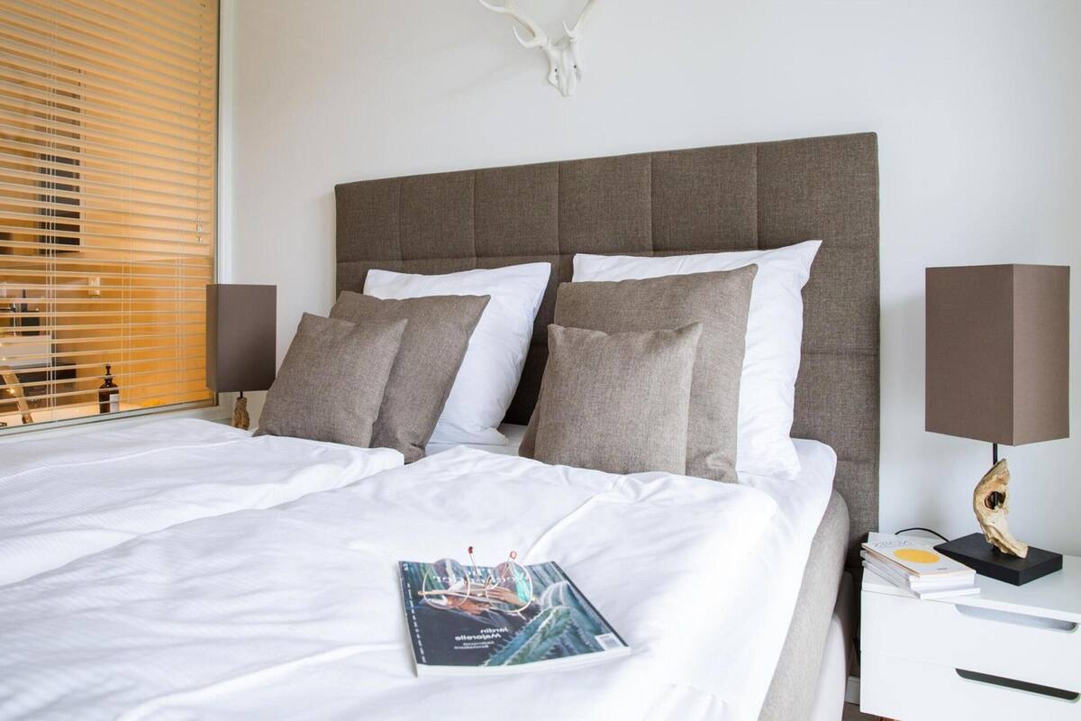 A neatly made bed with white and grey bedding, a magazine on the bed, and a wall-mounted deer skull decoration.