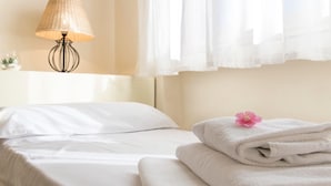 Een net opgemaakt bed met witte lakens, een lamp op een nachtkastje, en een raam met dunne gordijnen.