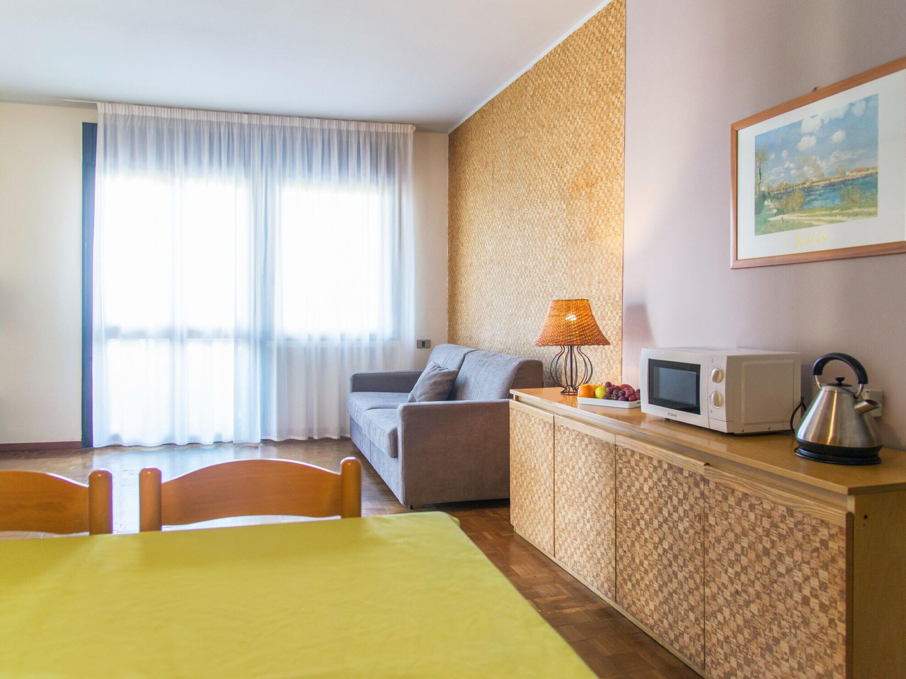 A hotel room with a bed, a sofa, a dining table, a microwave, a kettle, and a painting on the wall.