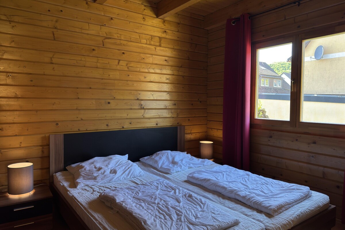 A wooden cabin room with a bed, a nightstand, a lamp, and a window with a view of houses.