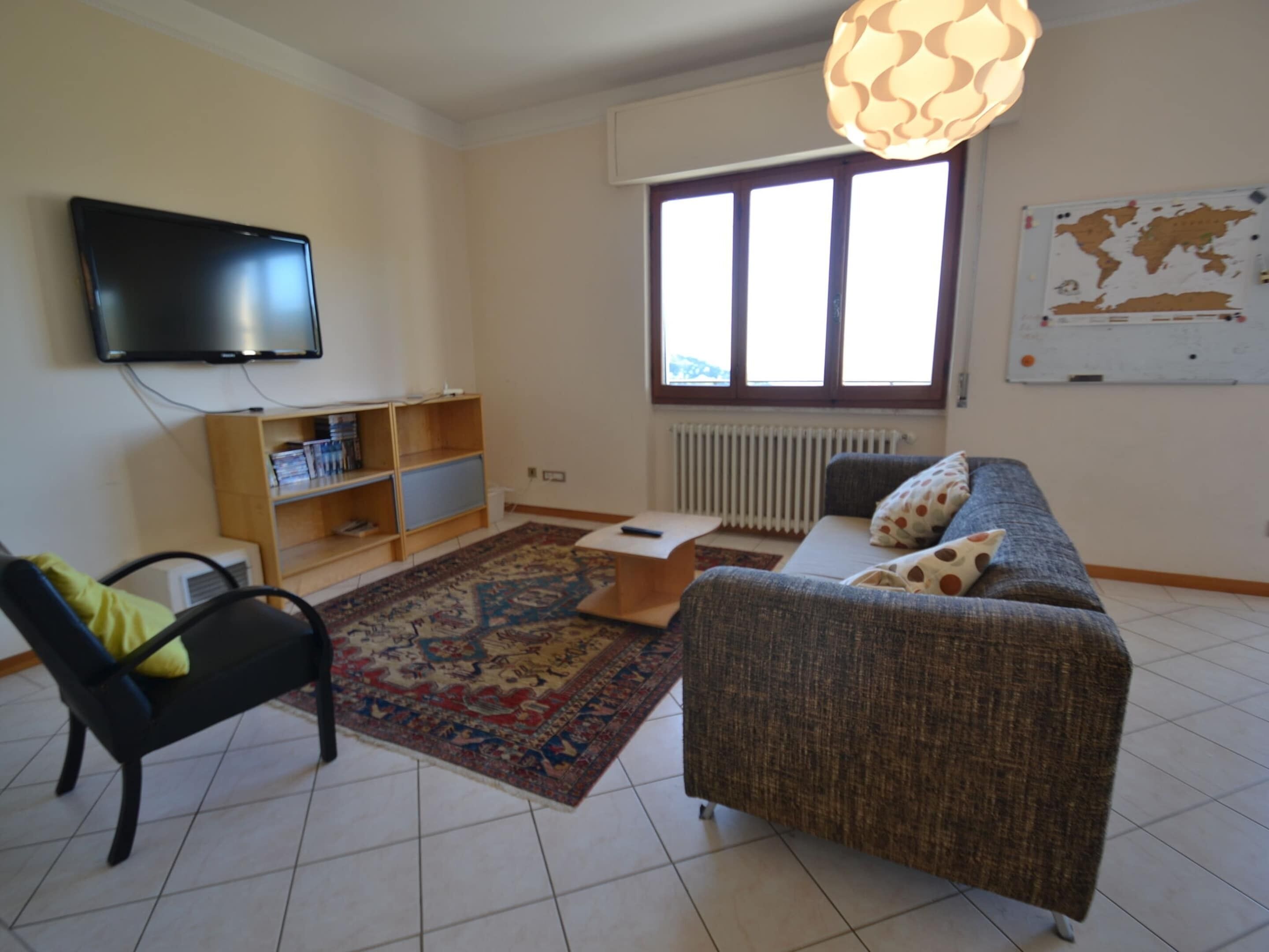 Una sala de estar con un sofá, una silla, un televisor, una estantería, una alfombra y una ventana con vistas al exterior.