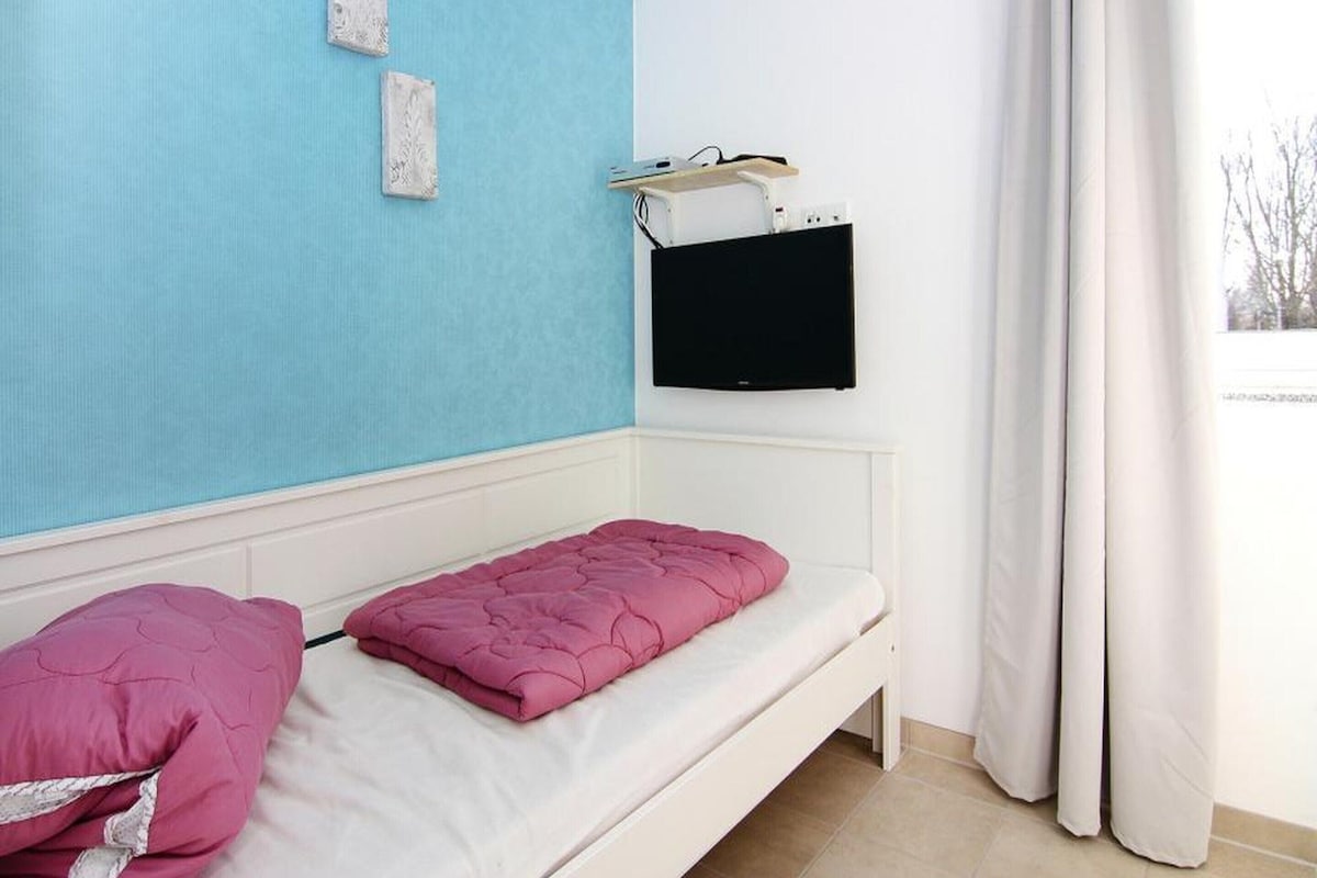 Ein Zimmer mit einem Einzelbett, einem an der Wand befestigten Fernseher und einem Fenster mit Vorhängen.