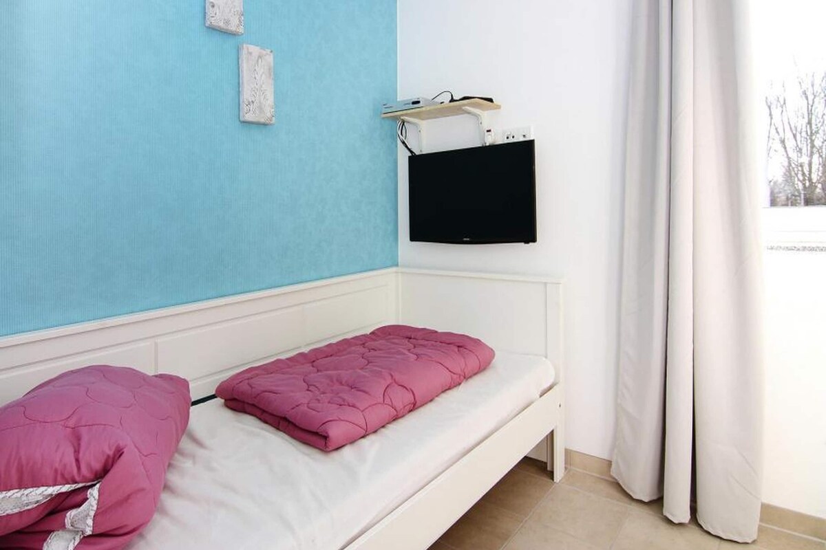 Une chambre avec un lit simple, un téléviseur fixé au mur et une fenêtre avec des rideaux.