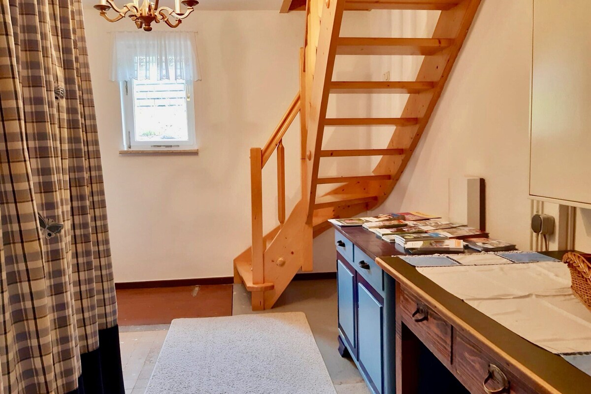 A room with a wooden staircase, a window with curtains, a desk with books, and a wardrobe.