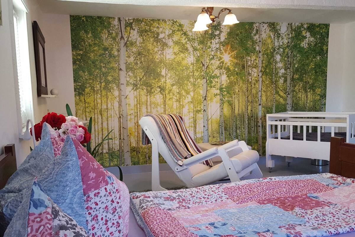 A baby room with a forest-themed wall mural, a white crib, a cushioned chair, and a bed with floral bedding.