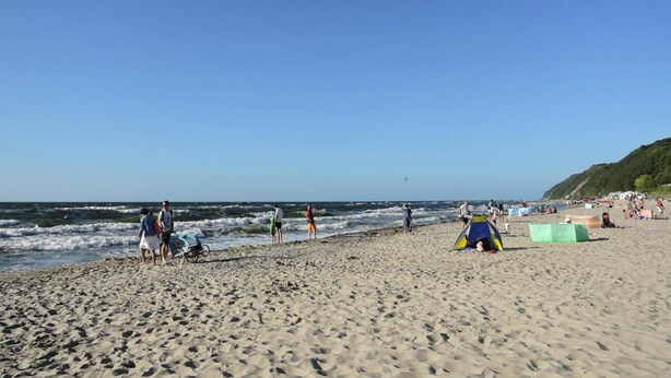 Une plage où l’on voit des gens se promener et se tenir près de l’eau, une tente installée, et un ciel dégagé.