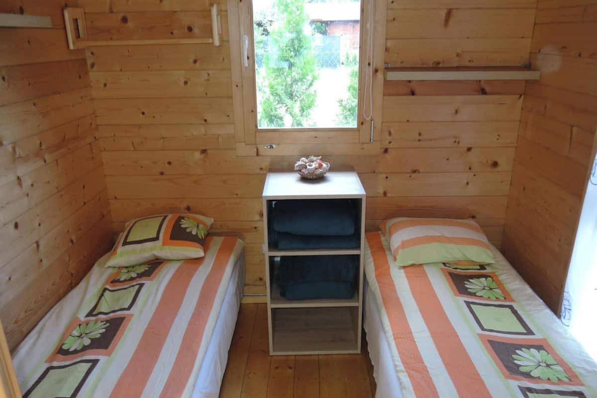 Um quarto de cabana pequeno e acolhedor, com duas camas de solteiro, uma prateleira de madeira com roupas dobradas e uma janela com vista para o verde lá fora.