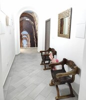 A hallway with a tiled floor, white walls, and a large arched doorway leading to an outdoor area. There are two wooden benches with cushions and a decorative mirror on the wall.