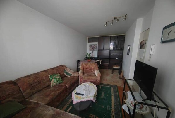 Una sala de estar con un sofá marrón, una mesita redonda con una jarrón, una silla, un televisor sobre un mueble bajo, y una cocina al fondo.