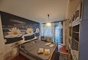 A bedroom with a bed, a large floral mural, a chandelier, and a view of the outside through a window with curtains. - Apartment Korzo (Bjelovar)