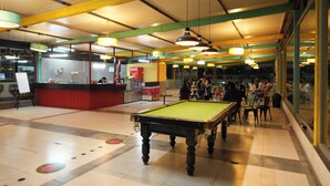 Une table de billard se trouve dans un espace intérieur bien éclairé, où l’on trouve également une table de billard américain, des sièges et un distributeur automatique de Coca-Cola.