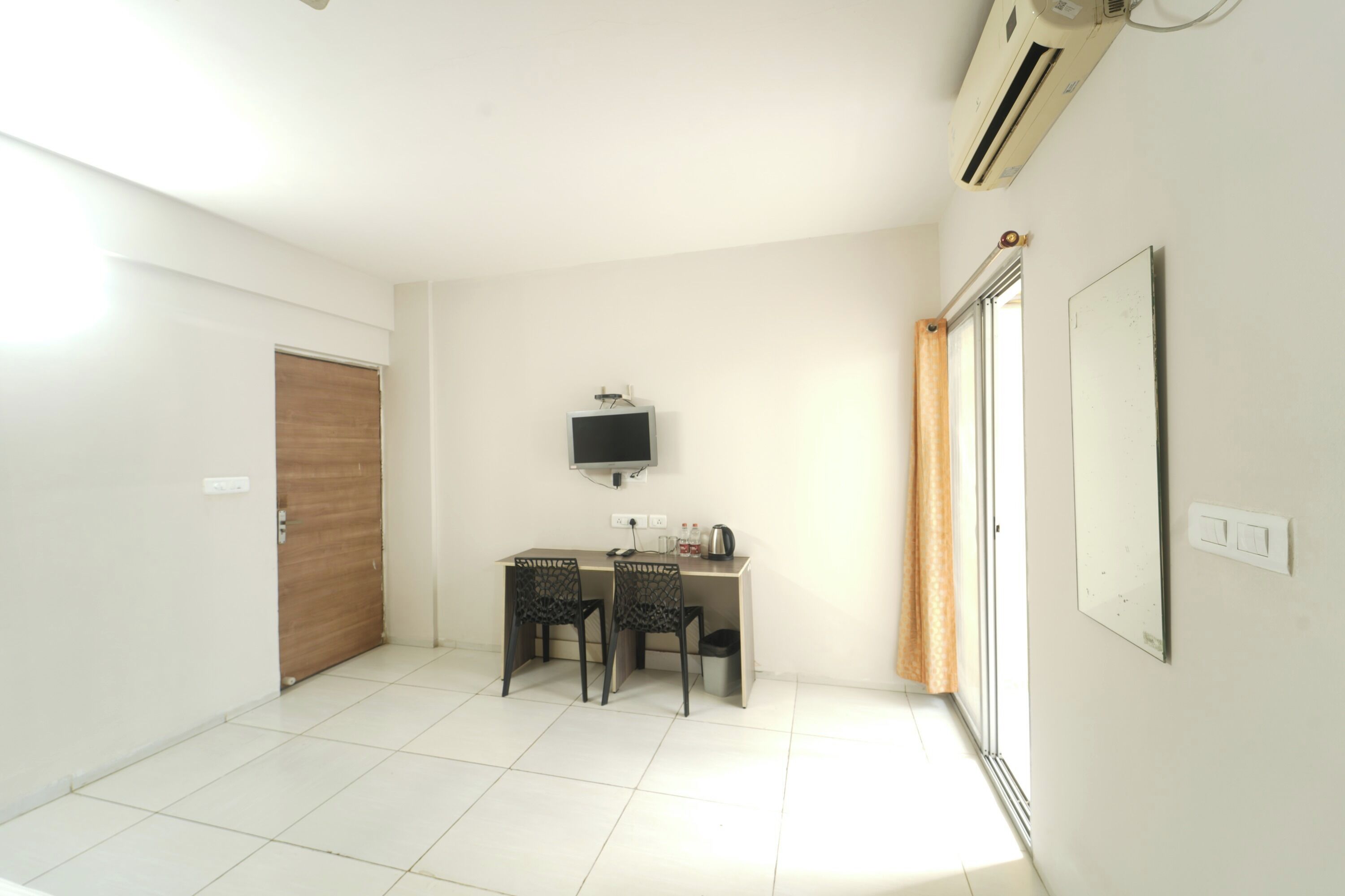 A small, bright room with a desk, two chairs, a television, and a window with curtains.