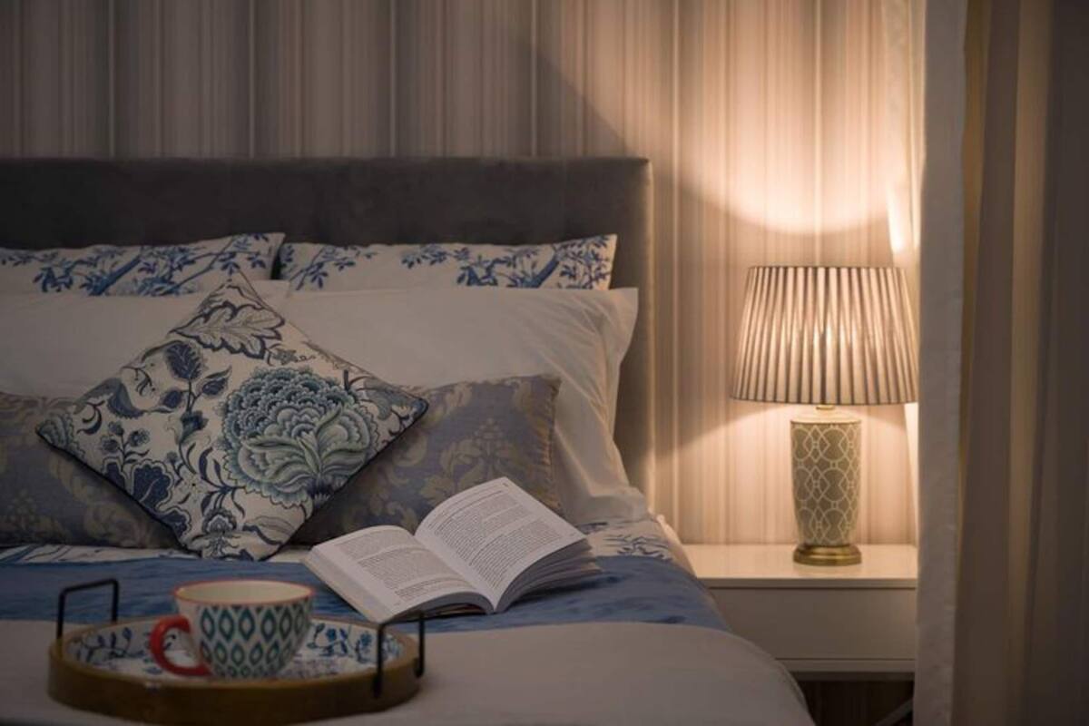 Un lit bien fait, agrémenté de coussins à motifs floraux bleus, une lampe de chevet et un plateau avec une tasse et un livre.
