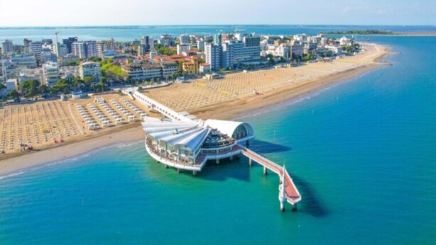 An aerial view of a coastal city with a prominent waterfront promenade, a large beach, and a distinctive modern building with a curved roof.