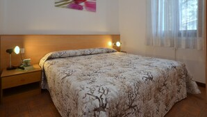 A bedroom with a bed, two bedside lamps, a nightstand, a wall-mounted picture, and a window with sheer curtains.