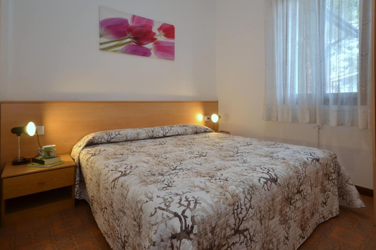 Un dormitorio con cama, dos lámparas de noche, una mesita de noche, un cuadro en la pared y una ventana con cortinas translúcidas.