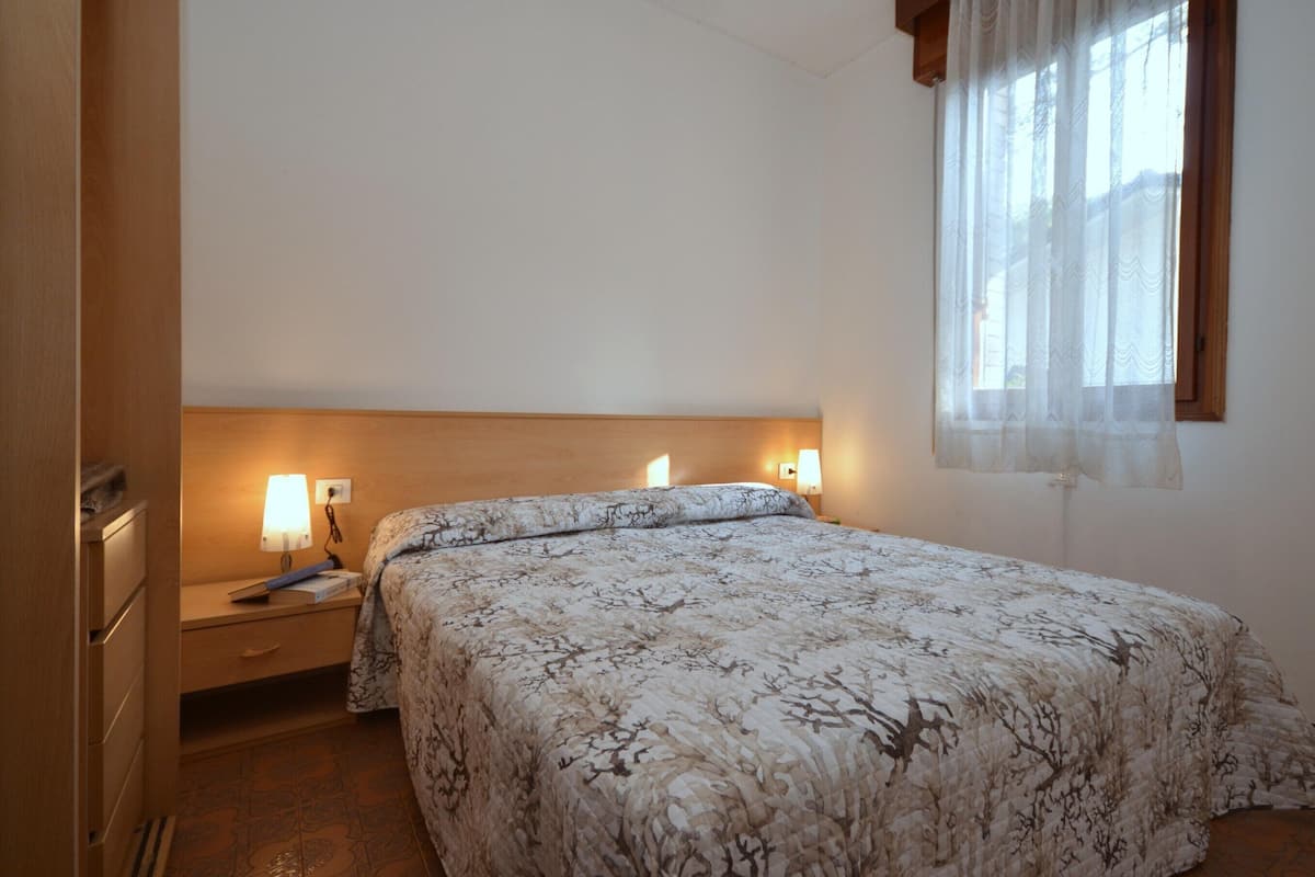 Un dormitorio con una cama, dos lámparas de noche, una mesita de noche y una ventana con cortinas translúcidas.