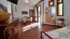 A room with a wooden dresser, a dining table with chairs, a kitchen area, and a bed with a patterned bedspread.